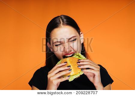 the girl eagerly eats an appetizing hamburger. the hungry girl eats. Isolated on an orange background.