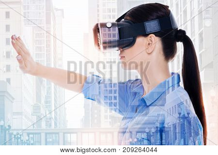 Immersed into VR. The side view of a pretty young woman using a VR headset and covering her sight with a hand while posing against an urban background
