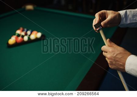 Caucasian Man wipes a cue with chalk ready to start game. Poolroom on the background.