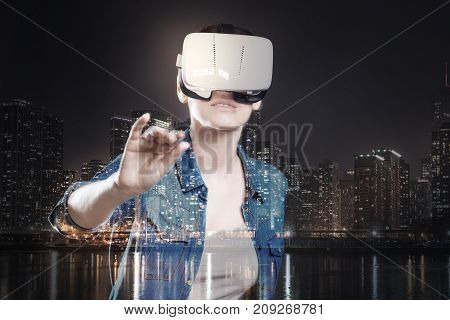 Getting closer. Pleasant young woman playing a VR game and putting her hand forward in a attempt to touch an object in front of her while standing against a night city background