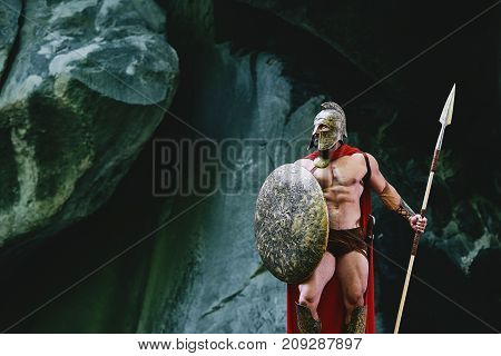 Epic shot of a medieval soldier wearing helmet and red cape holding a spear and a shield looking away thoughtfully copyspace.