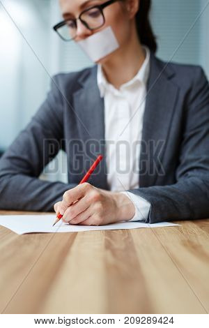 Young female with her mouth shut by cellotape making notes with pencil