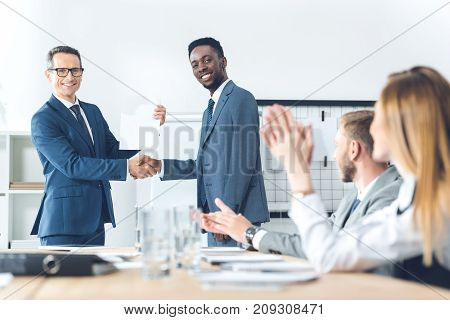 boss shaking hand of african american manager while colleagues clapping