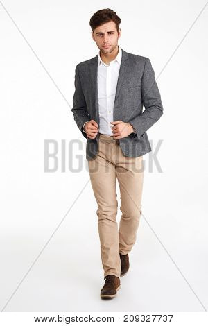 Full length portrait of a young handsome man in a jacket walking and looking at camera isolated over white background