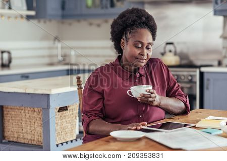 Smiling young African woman sitting alone at her kitchen table at home drinking coffee and working on a digital tablet