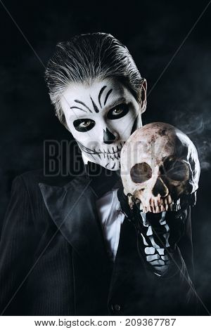Portrait of a child boy with carnival make-up of a skeleton wearing elegant tail-coat. Studio shot over black background. Halloween. Dia de los muertos. The Day of the Dead.