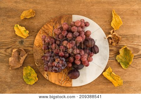 Upper view shot of grapes and plums on half-marble hald-wooden round board and autumn leaves around the plate and over wooden rustic background
