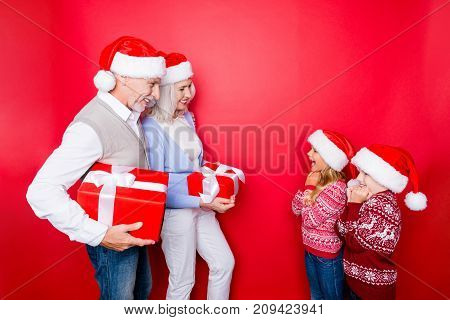 Four relatives: married senior couple of grandad and granny in knitted cute traditional x mas outfits prepard surprises for cheerful siblings full of emotions isolated on the red background