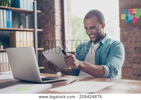 Cheerful Young Mulatto Worker Is Plying A Game On His Tablet In Office. He Is Very Excited, Wearing