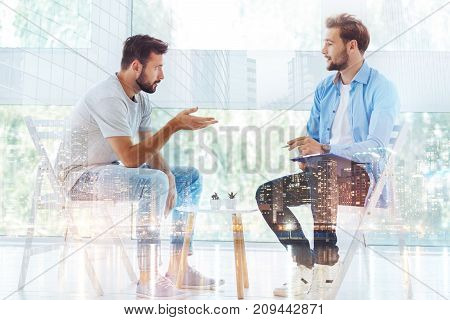 Being in trouble. Young handsome bearded man explaining his problems and raising right hand while psychologist making notes and listening to him carefully