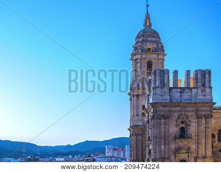 In the image we see the north and south towers of the cathedral of Malaga, behind them we see that there are buildings and mountains, all under a light blue sky.