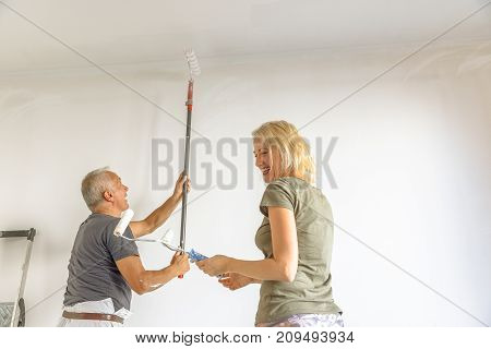 Smiling caucasian woman with senior painter man at work heaving fun whitewashing a white wall with paint roller in empty room for renovation.