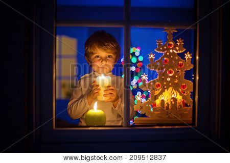 Beautiful little kid boy, standing by window at Christmas time and holding candle. Child waitng for Santa. With colorful lights from Christmas tree on background.