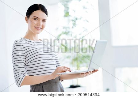 At work. Smart confident nice businesswoman standing against the window and smiling while using a laptop