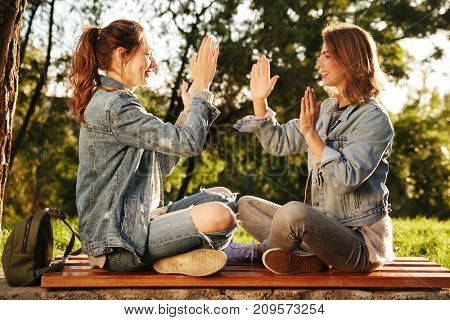 Two attractive young girls in casual jeans wear clap their hands while sitting on wooden bench in park outdoor