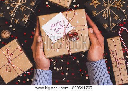 Man hands holding christmas holiday gift box with postcard merry xmas on decorated festive table with sparkle stars candy cane walnut on wooden background. Packaging gift wrap and ribbons twine. Winter time new year