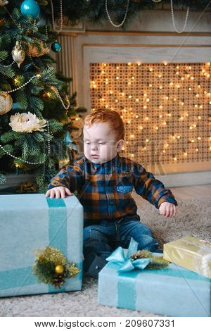 happy red-haired little boy sitting near boxes and opening Christmas presents near new year tree
