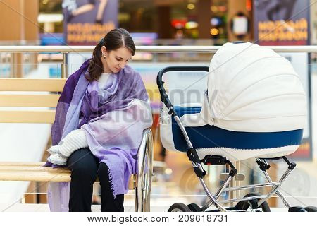 Young european woman with violet stole is breastfeeding her little child close to white baby carriage at public place shopping mall at day time