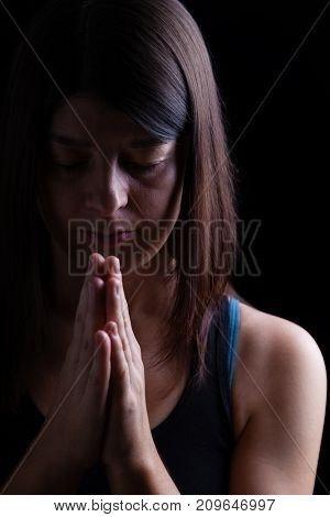 Faithful athletic woman praying, with hands folded in worship to god, head down and eyes closed in religious fervor, on black background. Concept for religion, faith, prayer and spirituality