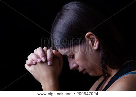 Faithful woman praying, hands folded in worship to god with head down and eyes closed in religious fervor, on a black background. Concept for religion, faith, prayer and spirituality.