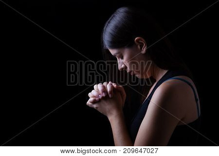 Faithful woman praying, hands folded in worship to god with head down and eyes closed in religious fervor, on a black background. Concept for religion, faith, prayer and spirituality.