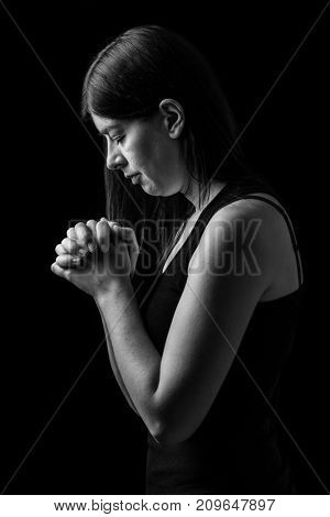 Faithful woman praying, hands folded in worship to god with head down and eyes closed in religious fervor, on a black background. Concept for religion, faith, prayer and spirituality.