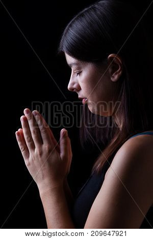 Faithful woman praying, with hands folded in worship to god, head down and eyes closed in religious fervor, on black background. Concept for religion, faith, prayer and spirituality