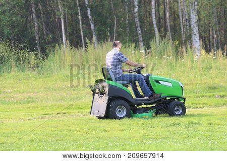 Lawn mower works on big green field near forest at summer day
