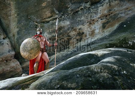 Full lengths shot of a Spartan warrior with sexy muscular strong athletic body standing near the rock wearing red cape holding a spear and a shield.