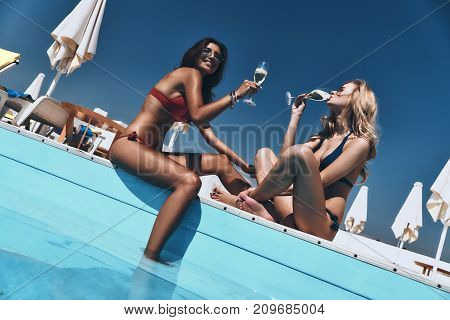 Toast to summer. Two stunning young women in bikini enjoying good drinks while sitting on the poolside outdoors