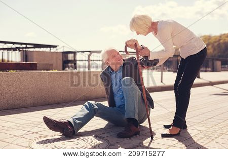 Giving support. Petite elderly woman holding her husbands arm and helping him to get up from the ground after falling down