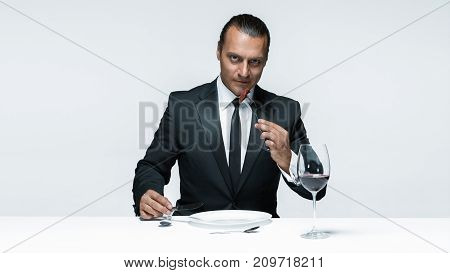 Portrait of a young man in image of maniak with knife, fork and and a piece of raw meat on a plate. Man in suit. The image in the style of Halloween