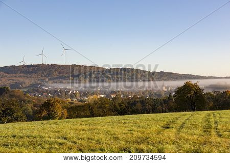 rural landscape grass field and trees in foreground fog mist over village in middle and hills and wind turbines in background against cloudless blue sky