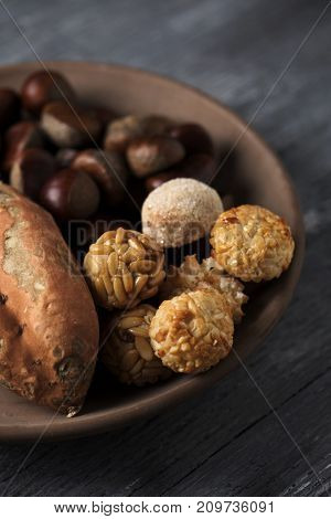 closeup of an earthenware bowl with some chestnuts, some panellets and a roasted sweet potato, as a typical snack eaten in All Saints Day in Catalonia, Spain, called castanada, on a wooden table