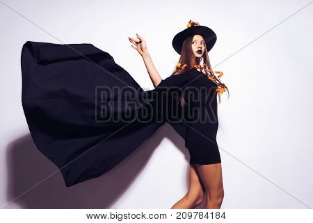 a young sexy gothic girl dressed in a witch for Halloween, standing in a big black hat, and a waving cloak