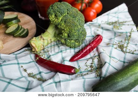 A large inflorescence of broccoli, a green squash and two red chili peppers are lying on the plaid tissue. in the background there are red tomatoes and a glass of tomato juice. Hurbs are used as decoration. nice