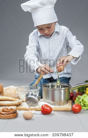 Cooking and Cuisine Concepts and Ideas.Portrait of Concentrated Caucasian Boy Mixing Fresh Egg in Saucepan. Posing In Cooking Hat Surrounded by Fresh Fruits and Vegetables. Against Gray. Vertical Image Orientation