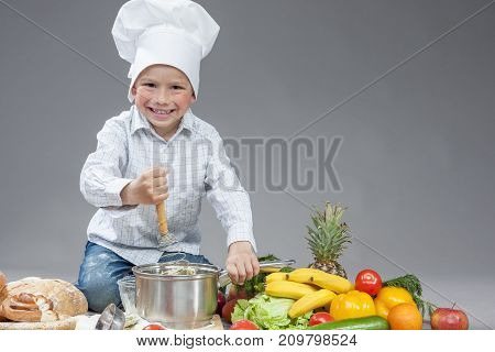 Portrait of Positive Smiling Caucasian Boy Working With Kitchen Ware In Cooking Hat. Surrounded by Fresh Fruits and Vegetables In Studio. Posing with Smiling Expression. Against Gray.Horizontal Image Orientation
