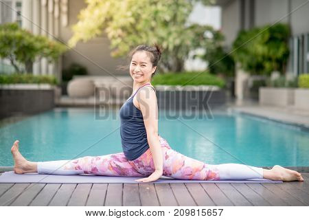 Beautiful young Asian woman doing yoga exercise with Hanumanasana posing (Monkey pose) near swimming pool. Healthy lifestyle and good wellness concepts