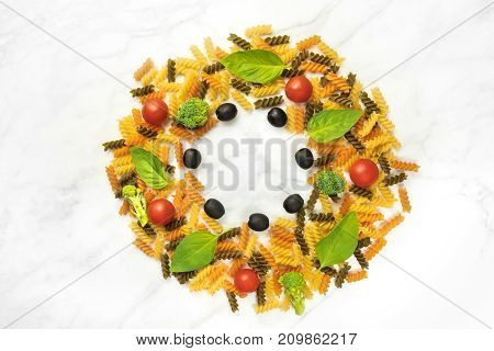 Pasta salad ingredients on white marble with a place for text. Rotini, cherry tomatoes, fresh basil leaves, and black olives, forming a circular frame for copy space