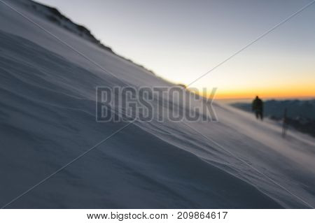 The blurred mountaineer professionally equipped at dawn of the sun walks on the slope in the early morning and the snow drifts over the snow above the snow. The concept of travel and conquest of steep peaks
