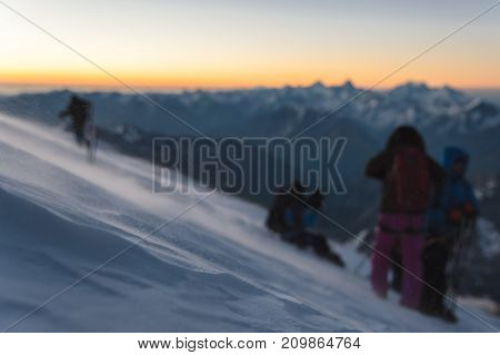 The blurred mountaineer professionally equipped at dawn of the sun walks on the slope in the early morning and the snow drifts over the snow above the snow. The concept of travel and conquest of steep peaks
