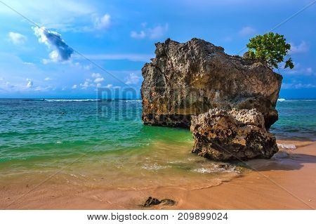 A large stone rock on the shore of the Bali Sea against the background of a surfer on a wave in sunny weather. Padang Padang Beach, Bali, Indonesia.