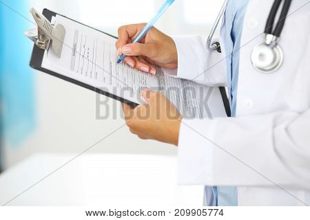 Close-up of a female doctor filling  up medical form at clipboard while standing straight in hospital.