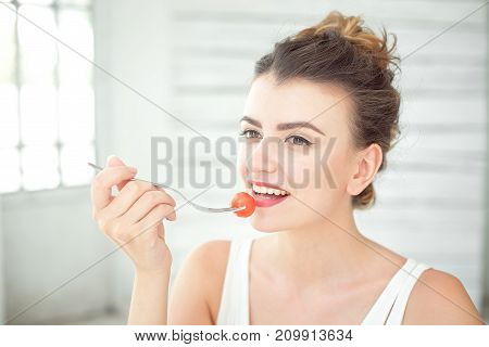 Close up portrait of attractive healthy young woman eating green salad indoors holding fork with fresh cherry tomato.
