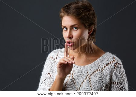 A dark-haired beautiful girl with short hair on a black background attached an index patsy near her mouth. Showing gesture quieter. Close-up.