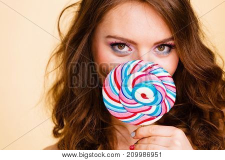 Woman attractive girl colorful eyes make up holding big lollipop candy in hand covering her face. Sweet food and enjoying concept. Studio shot on bright