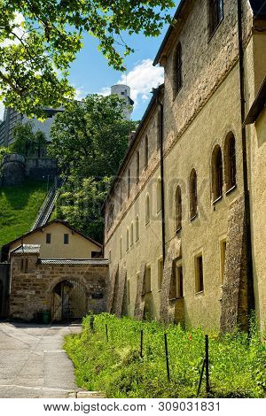 Old Church Wall And Path Beneath The Fortress Hohensalzburg On The Hill Above In Salzburg, Austria