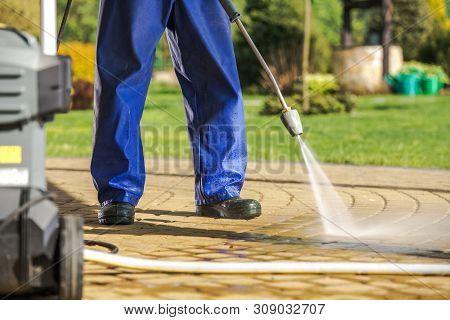 Worker And Pressure Washer. Men Wearing Rain Coat Cleaning Brick Paths Using Powerful Water Sprayer.
