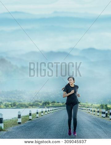 Young Woman Is Running On Road In The Morning With Mountains Natural Background.